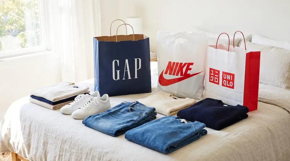 Свитеры, джинсы и кеды на кровати, рядом пакеты GAP, Nike и UNIQLO